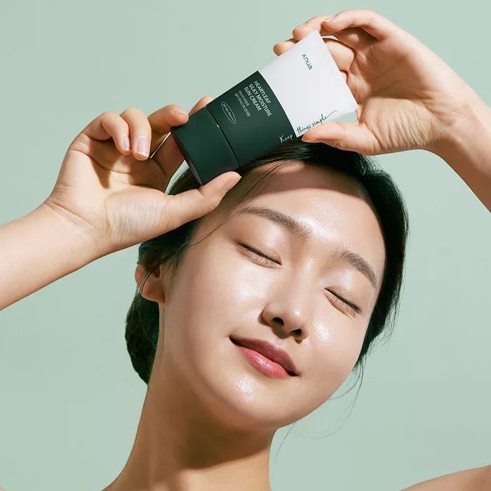 Skin TOGO model holding Anua Heartleaf Silky Moisture Sun Cream SPF50+ against a soft green background