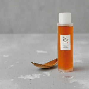 Skin TOGO Beauty of Joseon Ginseng Essence Water in a clear bottle with amber liquid and a wooden spoon on grey stone
