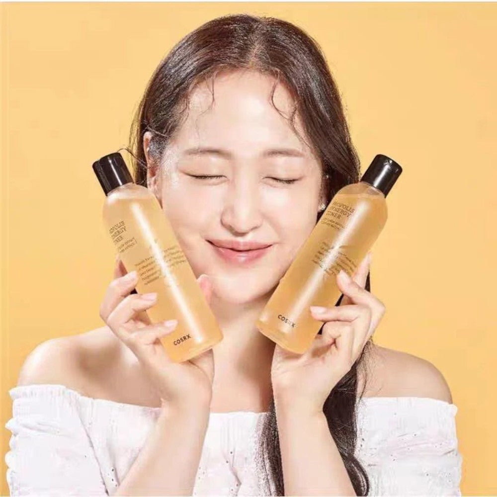 Skin TOGO model holding two bottles of COSRX Full Fit Propolis Synergy Toner against a soft yellow background.