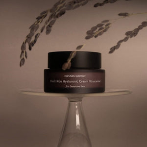 Skin TOGO Haruharu WONDER Black Rice Hyaluronic Cream in a dark jar on a glass pedestal with decorative dried plants.