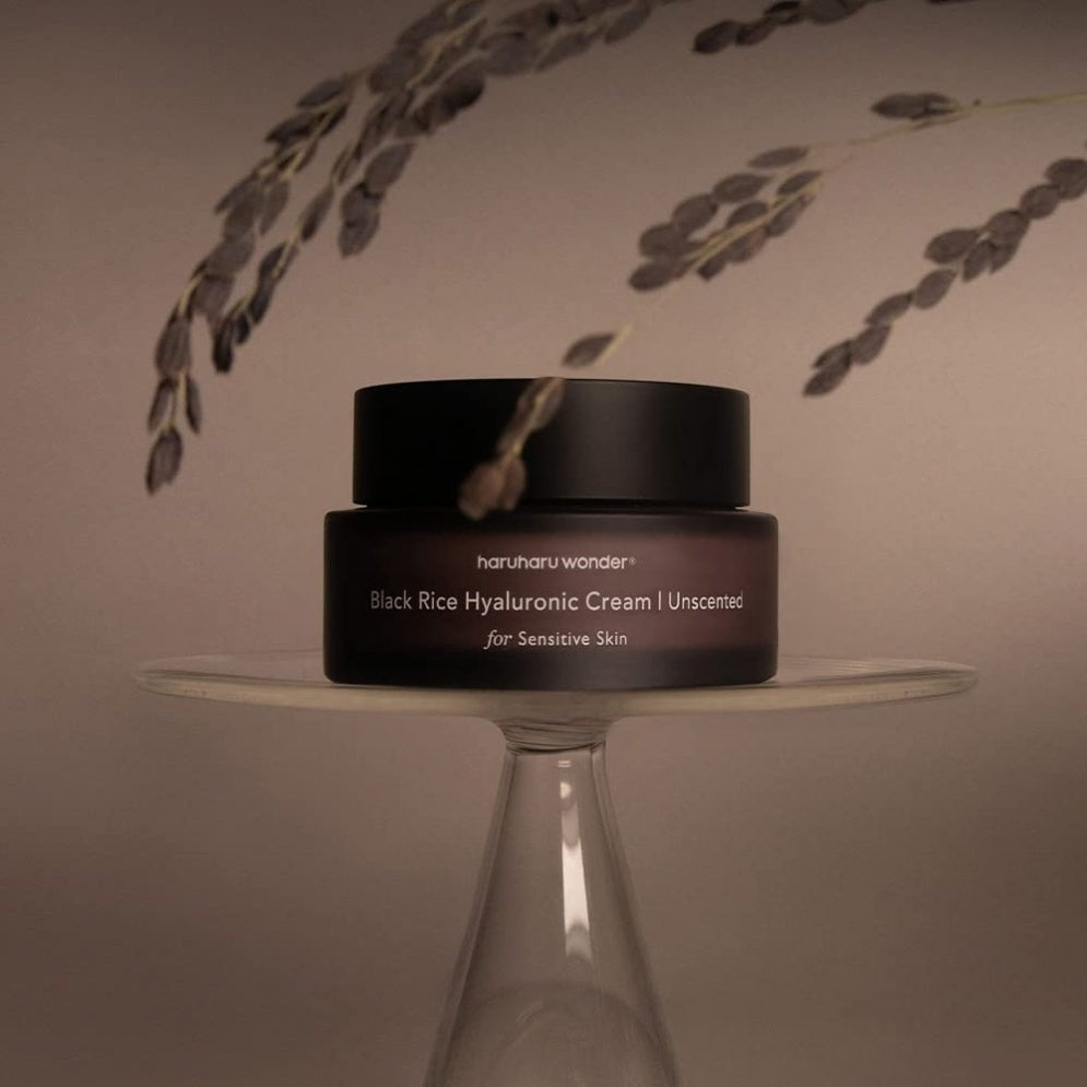 Skin TOGO Haruharu WONDER Black Rice Hyaluronic Cream in a dark jar on a glass pedestal with decorative dried plants.
