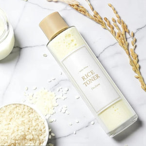 Skin TOGO I'm from Rice Toner in a clear bottle with milky liquid, surrounded by raw rice grains on a marble surface.