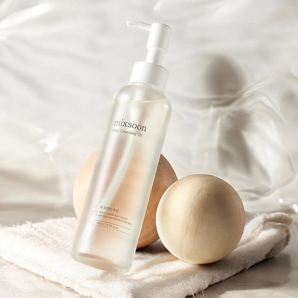 Skin TOGO Mixsoon Bean Cleansing Oil in a clear pump bottle with wooden spheres and a soft cloth on a white surface.