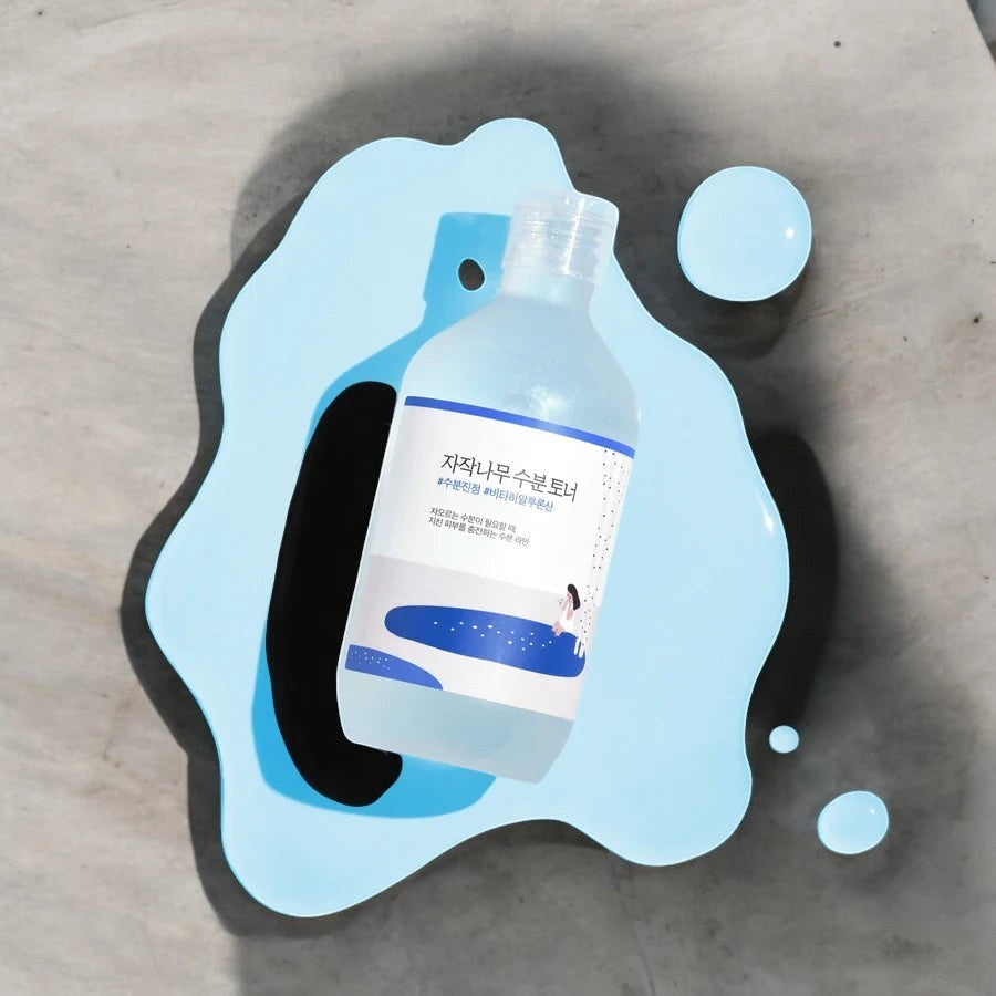 Skin TOGO Round Lab Birch Juice Moisturizing Toner bottle resting on a light blue liquid splash on a grey surface.