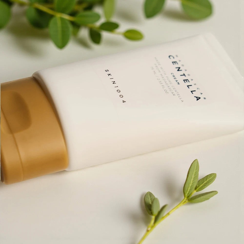 Skin TOGO SKIN1004 Madagascar Centella Cream in a white tube with a gold cap, surrounded by green leaves.