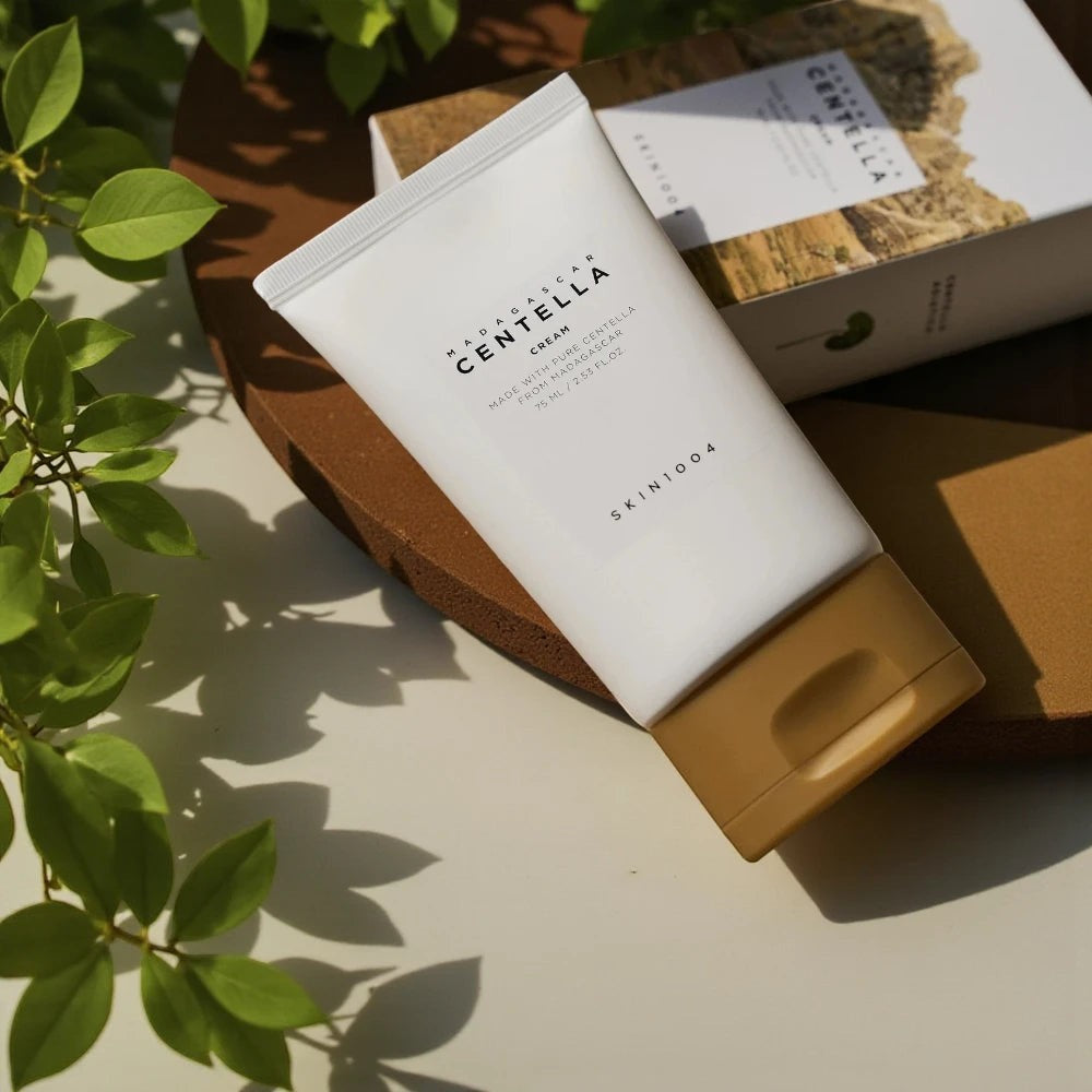 Skin TOGO SKIN1004 Madagascar Centella Cream in a white tube with a tan cap, placed on a brown surface with greenery.